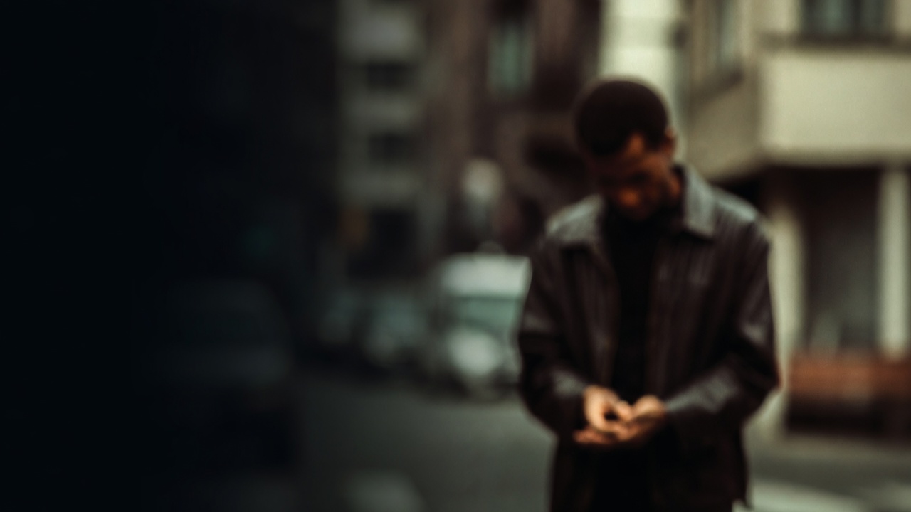 A softly blurred figure in a dark jacket stands on a city sidewalk, hands together